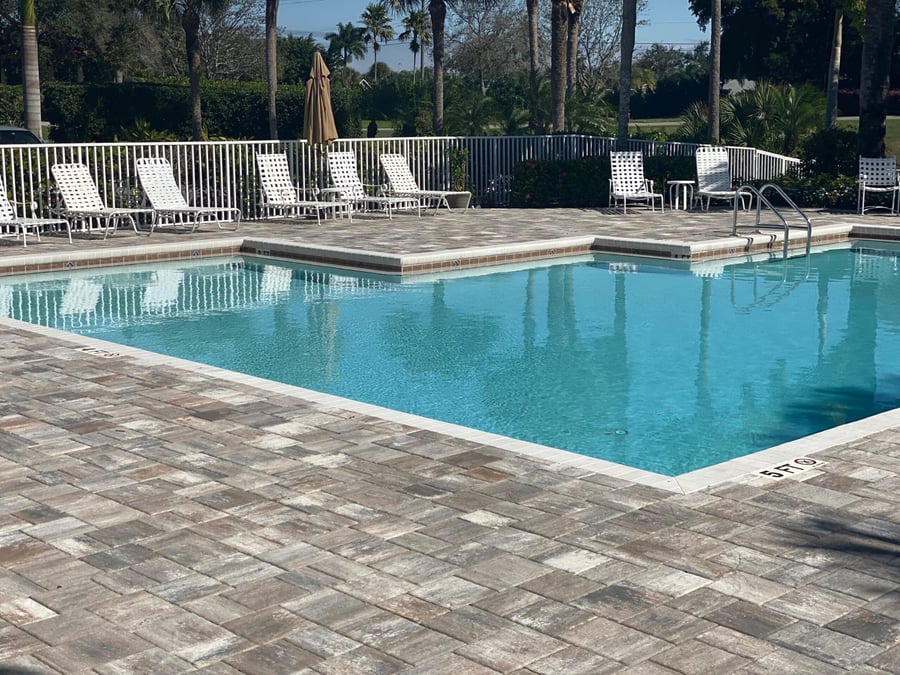Community pool deck in driftwood-toned pavers with white loungers
