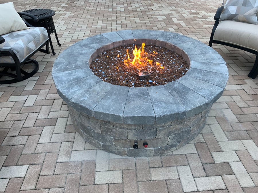 Round gas fire pit with fire glass on a herringbone paver patio