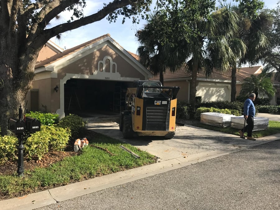 Skid steer removing an old villa driveway with paver pallets staged nearby